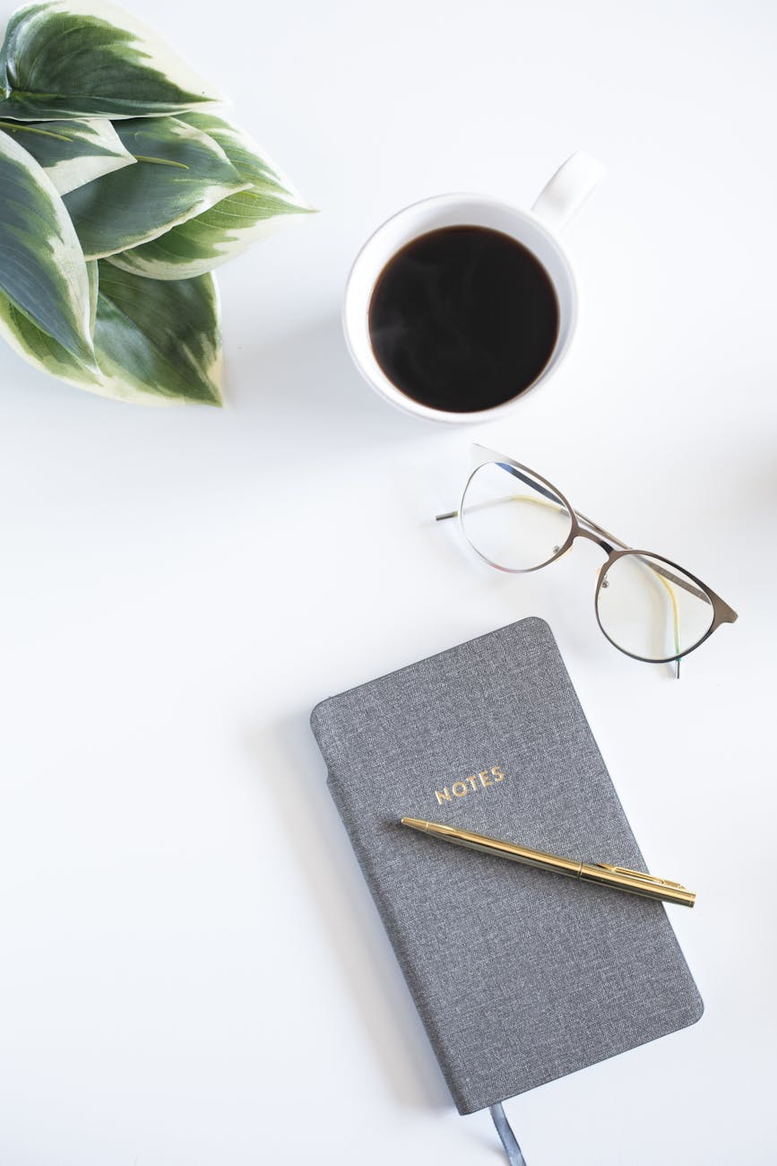 a notebook with pen near the eyeglasses and cup of coffee on white surface