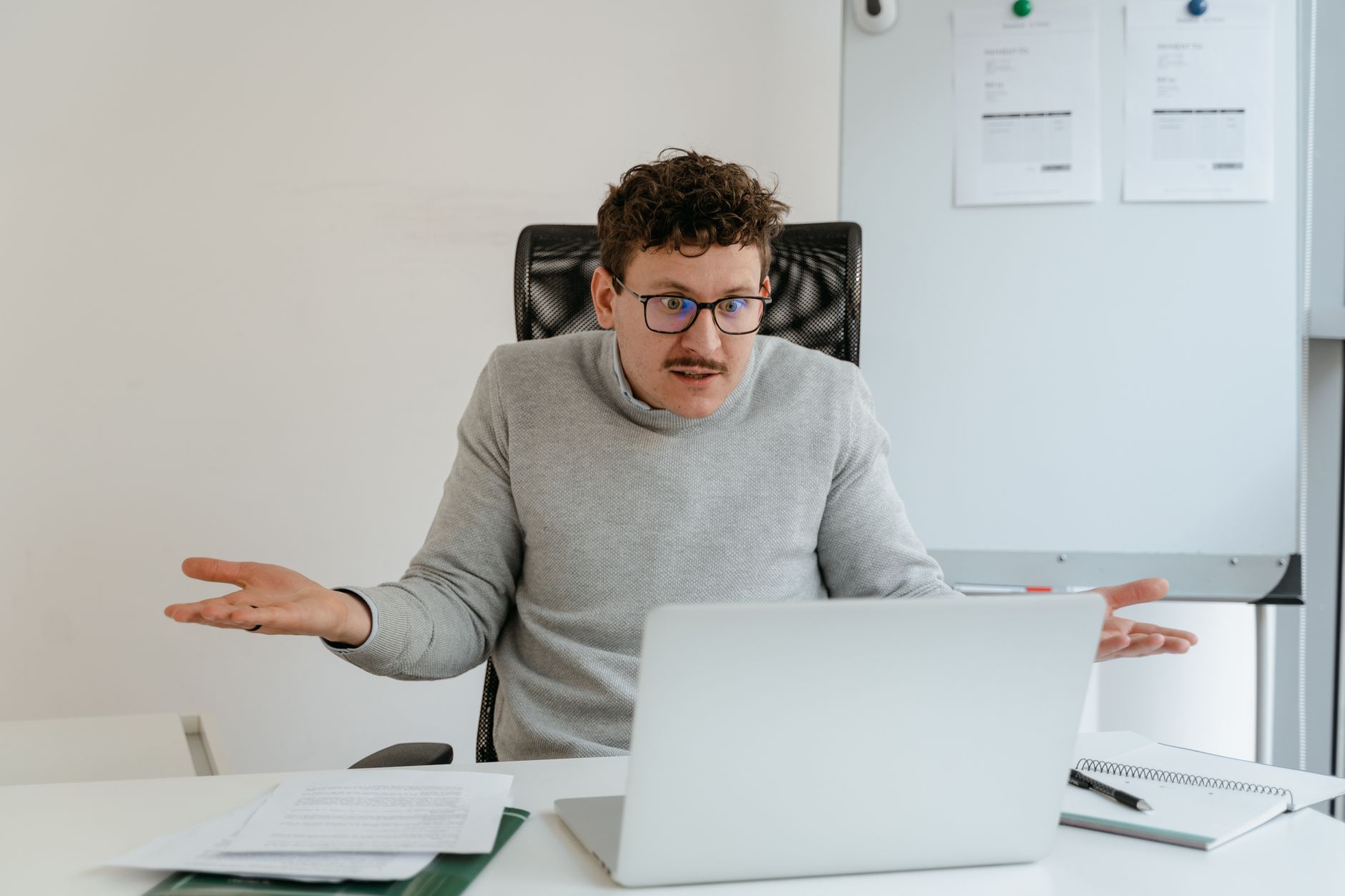 A man with glasses and a mustache sits at a desk in an office, shrugging with both hands raised while looking at a laptop. Papers and a notebook are spread on the desk, and a whiteboard is visible in the background.