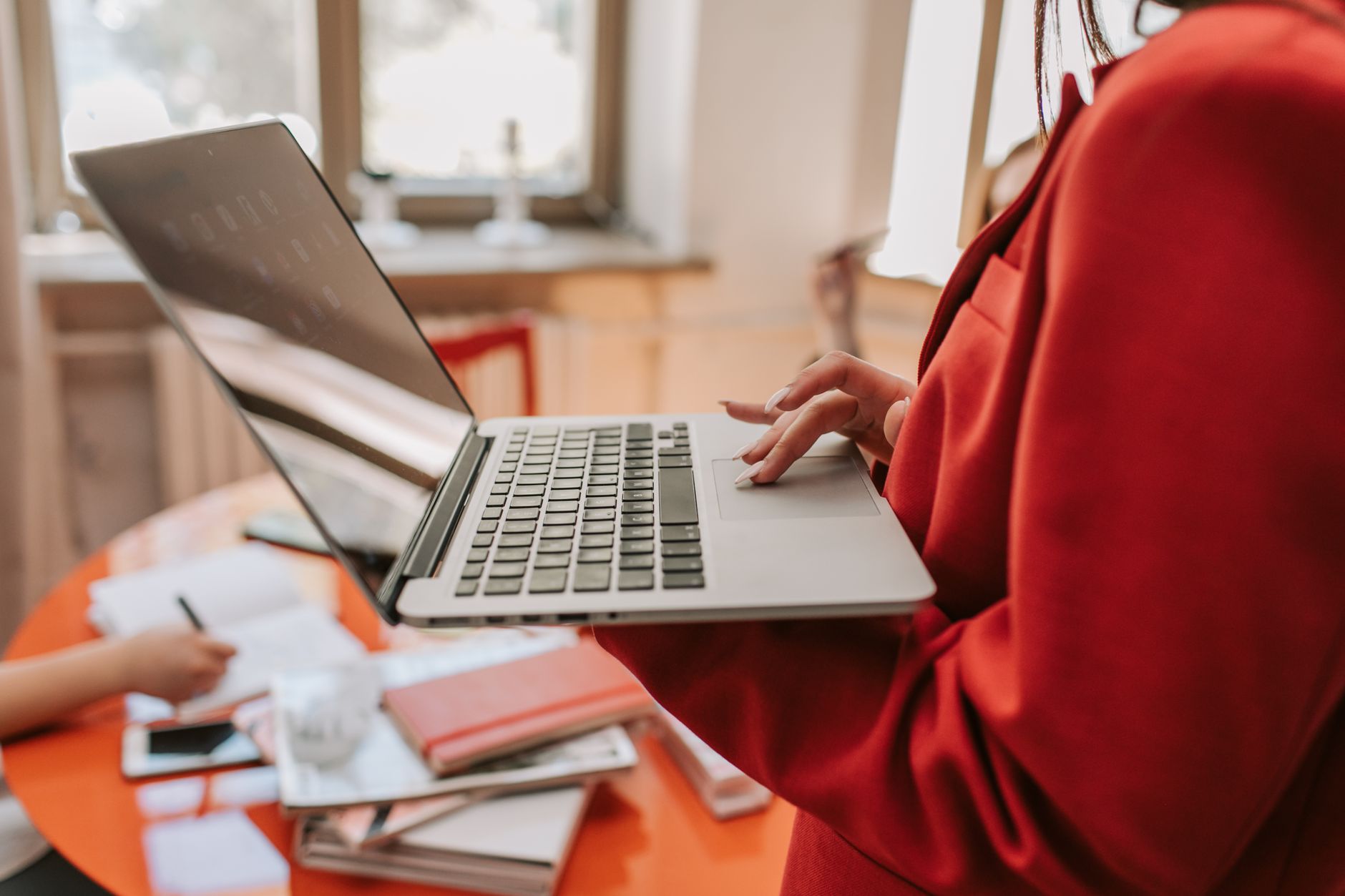 Close-up of a person in a bright red blazer holding an open laptop and using the trackpad, with a blurred office workspace and orange table in the background.